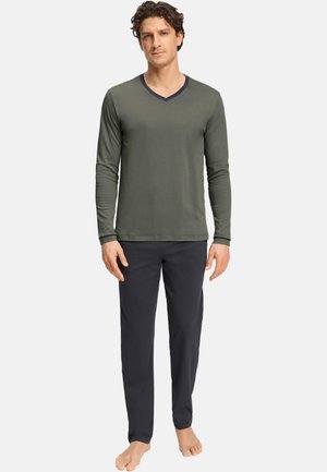 Mann steht barfuß und trägt ein olivgrünes V-Ausschnitt-Shirt mit langen Ärmeln und eine schwarze Hose vor weißem Hintergrund.