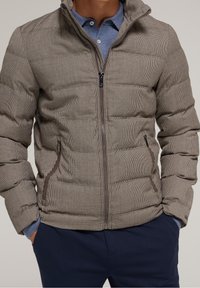 Chaqueta acolchada de patrón a cuadros marrones, con cierre de cremallera, cuello alto y bolsillos laterales. El material parece suave y texturizado.