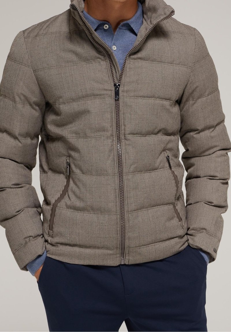 Chaqueta acolchada de patrón a cuadros marrones, con cierre de cremallera, cuello alto y bolsillos laterales. El material parece suave y texturizado.