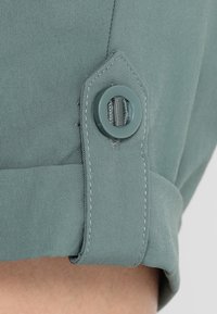 Dämpad teal-tyg med en slät yta, med en rund knapp och lösa sömmar på ärmslutet.