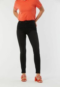 Jean skinny taille haute noirs associés à un t-shirt orange et des sandales à talons carrés rouges, mettant en valeur un design ajusté et une texture lisse.