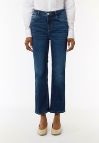 Dunkelblaue Jeans mit geradem Bein, fünf Taschen-Design und kontrastierenden orangefarbenen Nähten. Getragen mit hellen Ballerinas.
