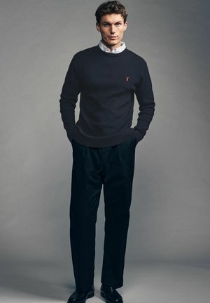 Pulover în navy închis cu textură canelată, având un mic logo roșu pe pieptul stâng; pantalonii negri și pantofii completează ținuta.