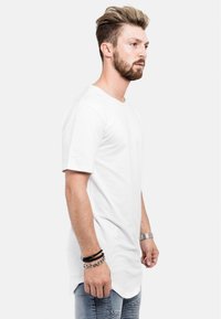 T-shirt en coton blanc à manches courtes, col rond et ourlet arrière plus long. Porté avec un jean en denim bleu présentant des détails texturés.