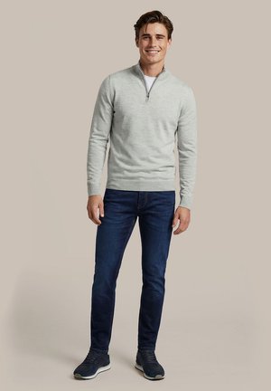 Lachende man staand met een lichtgrijze sweater met rits bij de hals, donkerblauwe spijkerbroek en marineblauwe sneakers op een neutrale beige achtergrond.