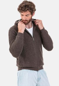 Sweat à capuche marron à fermeture éclair, fabriqué en tissu texturé avec une finition douce, doté d'une capuche et de poignets côtelés. Porté sur une chemise blanche.