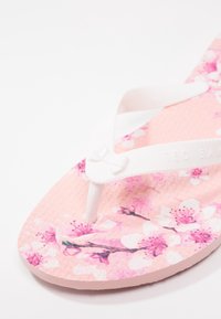 Tongs roses avec des brides blanches, détail de nœud en relief et un motif floral de fleurs de cerisier sur la semelle.