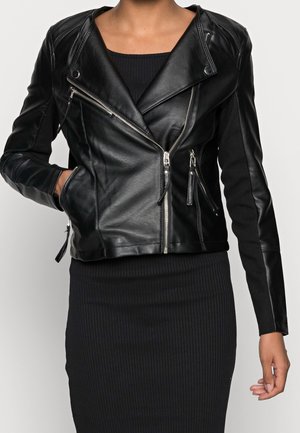 Frau trägt eine schwarze Leder-Bikerjacke mit silbernen Reißverschlüssen über einem schwarzen, gerippten Kleid, eine Hand in der Jackentasche.