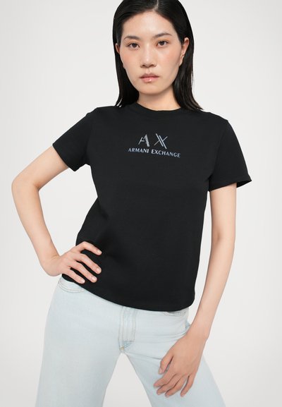 T-shirt en coton noir arborant le logo "AX ARMANI EXCHANGE" en argent. Manches courtes, col rond et coupe décontractée. Associé à un jean bleu clair.