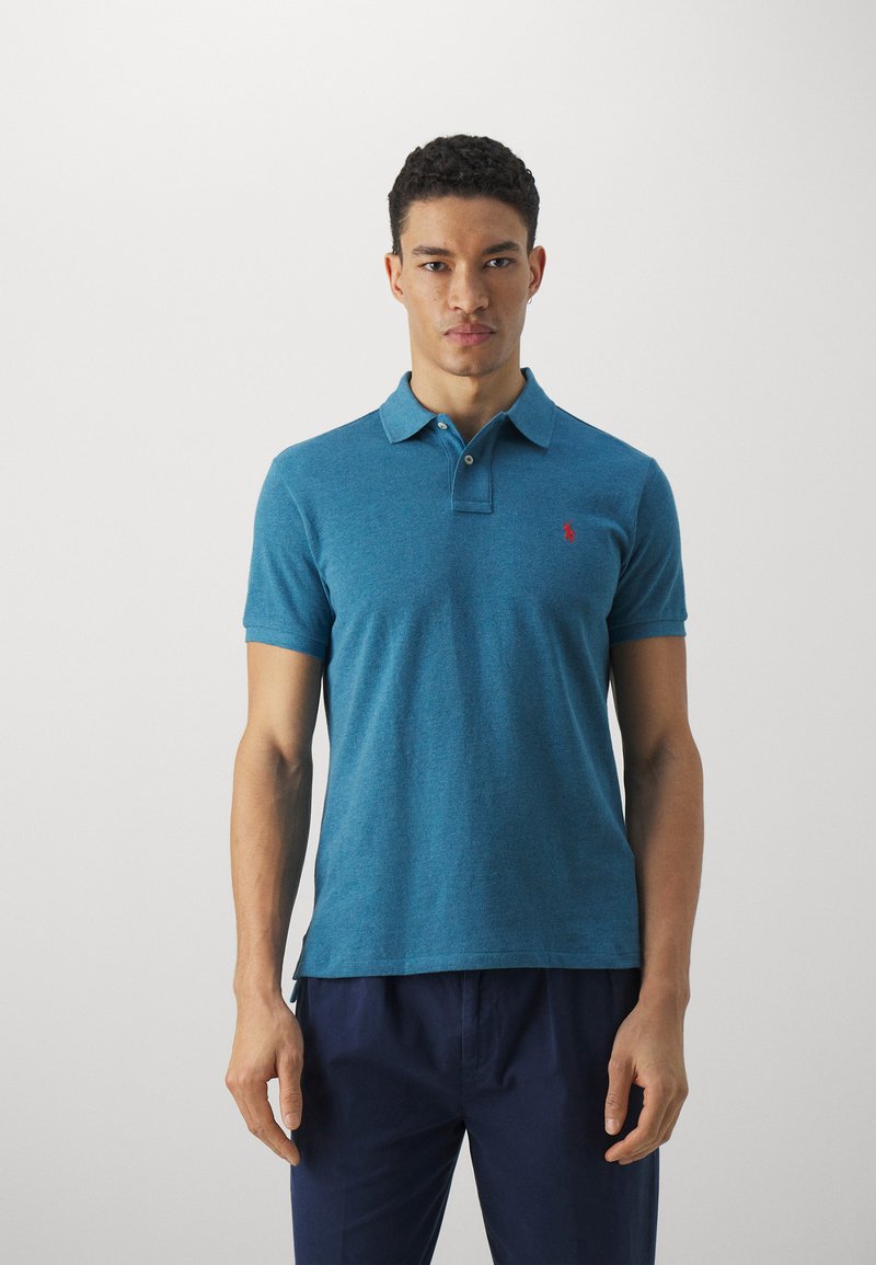 Polo en tejido azul pizarra, de manga corta, con cuello y un pequeño logo rojo en el pecho. Corte ajustado, textura suave.