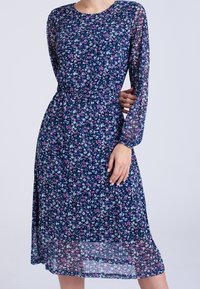 Kukkakuvioinen midi-mekko, jossa on pitkät läpinäkyvät hihat, tummansininen perusväri, hajallaan vaaleanpunaisia ja valkoisia kukkia, vyötäröltä pussittava leikkaus ja leikkisä helma.