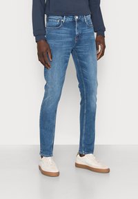 Vaqueros de mezclilla azul con un corte ajustado, cinco bolsillos y costuras en contraste. Combinados con zapatillas blancas y una camiseta de manga larga azul marino.