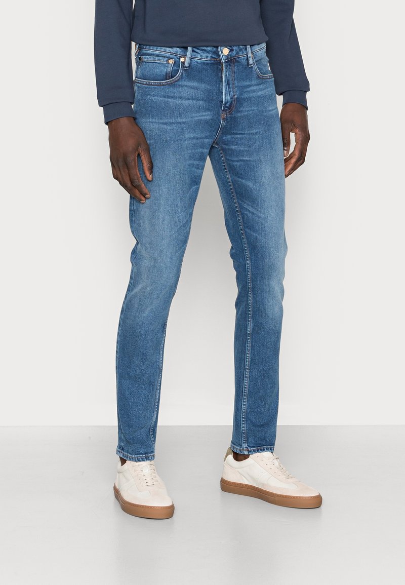 Vaqueros de mezclilla azul con un corte ajustado, cinco bolsillos y costuras en contraste. Combinados con zapatillas blancas y una camiseta de manga larga azul marino.