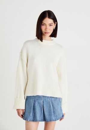 Ecrufarbener Strickpullover mit geripptem Kragen und Bündchen, kombiniert mit einem blauen Jeansrock mit Falten und fransigem Saum.