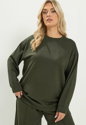 Blondynka ubrana w luźny, ciemnozielony top z długim rękawem i pasujące spodnie, stojąca na jasnym tle.