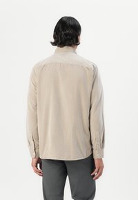 Camisa a rayas color beige con mangas largas, cuello abotonado y botones en los puños; presenta un corte holgado y una textura suave.