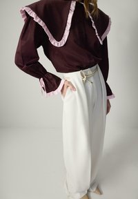 Bordeauxkleurige blouse met roze gerimpelde kraag en manchetten, gecombineerd met witte high-waisted broek, vastgemaakt met een beige strik, met taps toelopende benen.