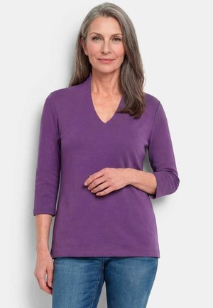 Femme aux cheveux gris portant un haut violet à manches trois-quarts en V et un jean bleu, debout devant un fond clair uni.