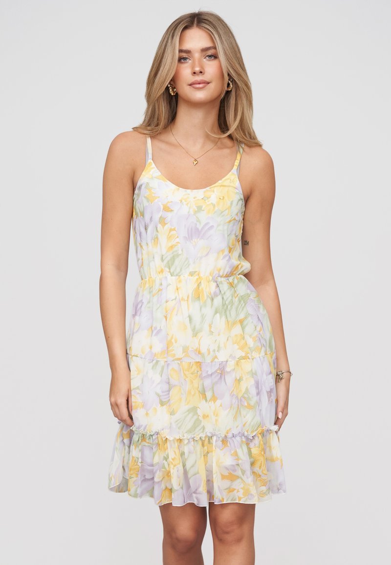 Jeune femme aux cheveux blonds portant une robe sans manches à volants floraux dans des teintes pastel jaune, vert et violet sur un fond uni.
