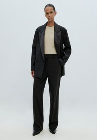 Zwarte double-breasted blazer over een beige aansluitend top, gecombineerd met zwarte op maat gemaakte broeken en puntige zwarte hakken. Gladde textuur overal.