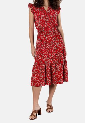 Femme portant une robe midi rouge à motifs floraux avec des manches courtes à volants et des sandales à talons marron, debout devant un fond uni.