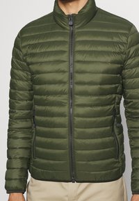 Giacca imbottita verde oliva con colletto alto, chiusura a zip e due tasche laterali. Presenta una trama leggera e liscia con cuciture orizzontali.