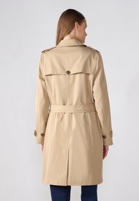Beige trench coat dengan detail belakang berbutang, pinggang berikat, dan epaulet bahu. Fabrik licin dan kalis air dengan siluet yang menyanjung.