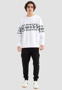 Sudadera de algodón blanca con texto negro en las mangas, combinada con pantalones cargo negros y zapatillas blancas con suelas beige. Diseño minimalista.