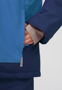 Giacca leggera blu e navy con una texture liscia, dotata di tasca laterale, polsini regolabili e etichetta con logo sull'orlo.