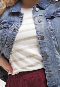 Jeansjacka med mörka knappar, bärs över en vit randig skjorta med guldiga ränder, lager ovanpå en vinröd kjol.