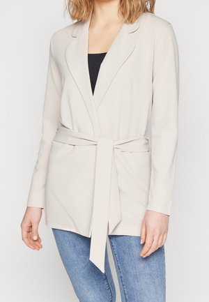 Femme portant un blazer beige clair ceinturé sur un haut noir et un jean bleu, debout devant un fond blanc uni.