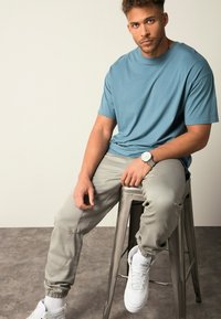 Lichtblauw T-shirt, grijze joggers, witte sneakers. Model zit op een metalen kruk met een horloge accent. Casual sportieve kleding.