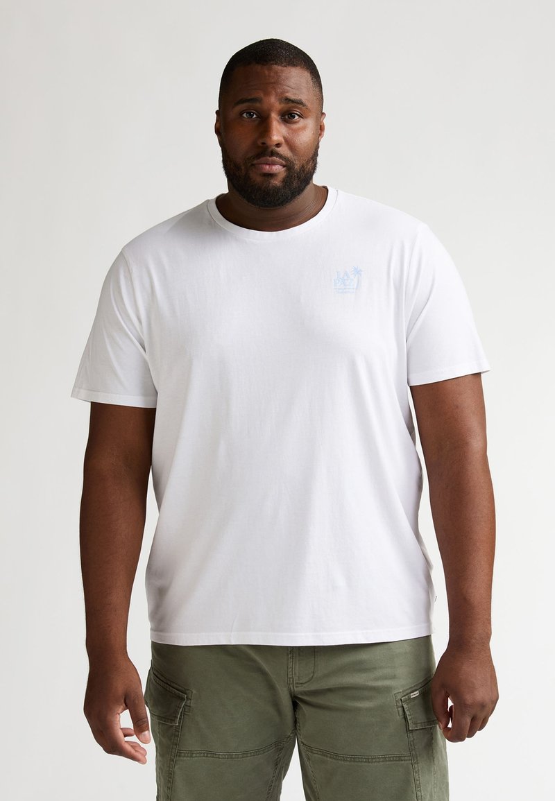 T-shirt en coton blanc avec col rond et manches courtes. Présente un détail de logo bleu clair sur la poitrine. Porté avec un pantalon cargo vert olive.