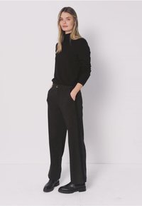 Maglione nero a collo alto abbinato a pantaloni a gamba larga di colore grigio scuro. La modella è in piedi con le mani nelle tasche, indossando scarpe nere slip-on.