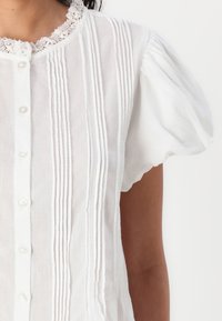 Hvit bluse med en blondekrage, korte puffede ermer og vertikale folder foran. Seks knapper gir funksjonell detalj.