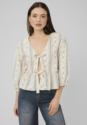 Femme portant un chemisier crème à rayures verticales noires, petits détails floraux orange, manches bouffantes et deux fermetures à nouer à l'avant, assorti à un jean bleu.