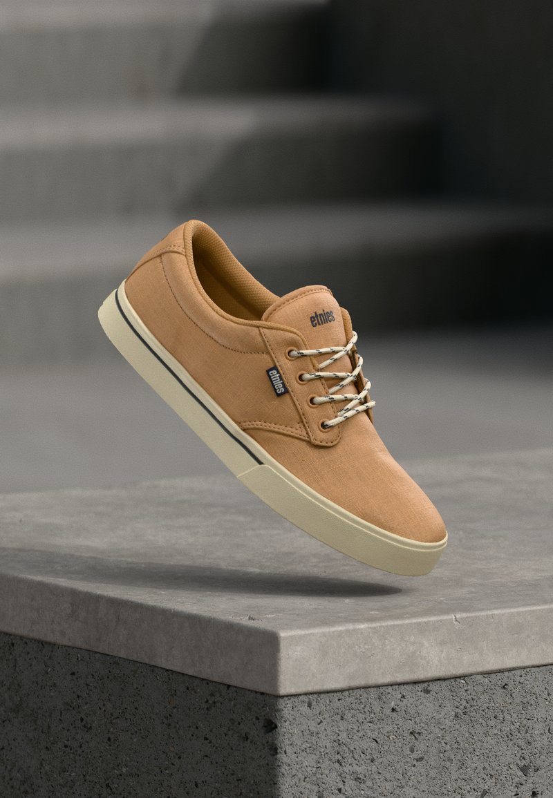 Baskets en toile couleur beige avec lacets blancs et semelle crème marquée "etnies", posées au-dessus d'un rebord en béton avec en arrière-plan des escaliers flous.
