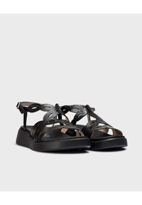 Las sandalias negras presentan una parte superior texturizada con correas entrelazadas y una suela plana. El diseño incluye una hebilla para el ajuste y es de punta abierta.