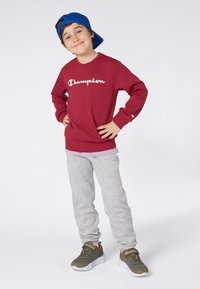 Gutt som har på seg en blå caps bak-fram, rød Champion sweatshirt, grå joggebukser og grønne joggesko, står selvsikkert med hendene på hoftene.
