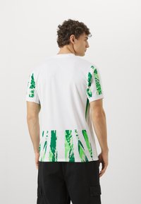 Chemise à manches courtes blanche avec motif abstrait vert sur les manches et le bas du dos. Fabriquée en tissu léger avec une texture douce.