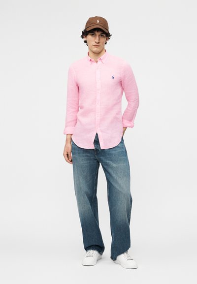 Jauns vīrietis ar brūnu cepuri, gaiši rozā kreklu ar pogām, vaļīgām zilajām džinsu biksēm un baltām sporta kurpēm stāv pret vienkāršu fonu.