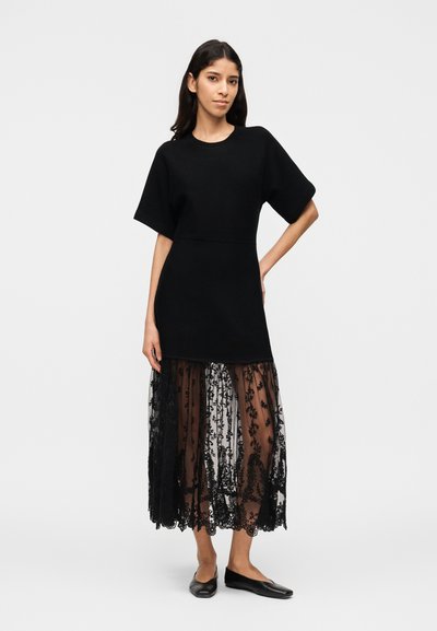 Γυναίκα στέκεται φορώντας μαύρο φόρεμα με κοντά μανίκια και διάφανο δαντελένιο στρώμα στη φούστα, συνδυασμένο με μαύρα επίπεδα παπούτσια, μπροστά σε απλό φωτεινό φόντο.