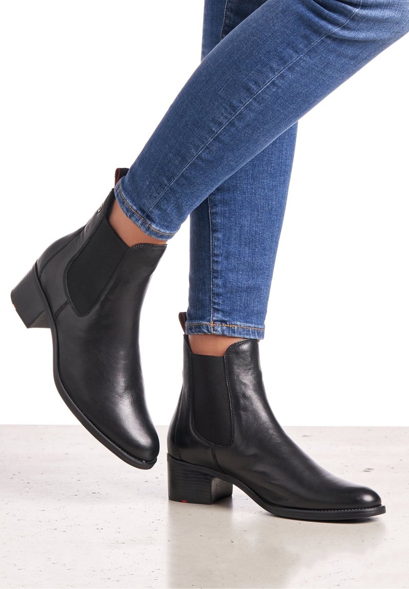 Bottes cheville en cuir noir avec des talons carrés bas, portées avec un jean skinny bleu sur un fond de sol clair.