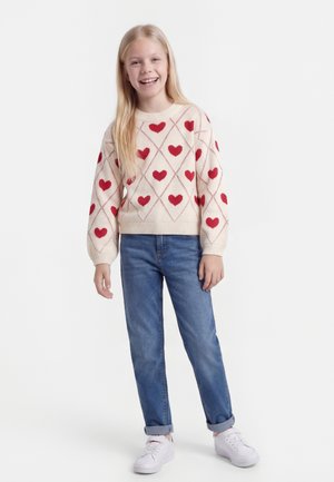 Fille souriante aux longs cheveux blonds portant un pull blanc avec des cœurs rouges, un jean bleu et des baskets blanches, debout devant un fond blanc.