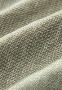 Texturovaná tkanina v olivově zeleném a krémovém herringbone vzoru, která ukazuje jemnou, tkanou texturu a podrobné diagonální linie.