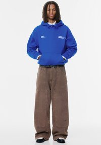 Felpa blu con cappuccio, dotata di tasca frontale e stampa del logo; abbinata a pantaloni larghi marroni, con cuciture visibili e tasche laterali.