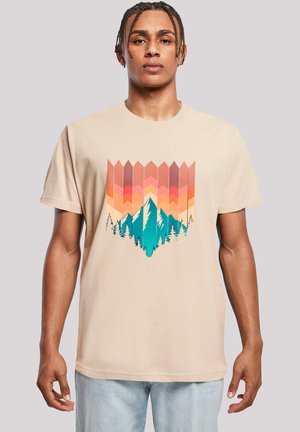 Young man with braided hair wearing a beige t-shirt featuring a colorful geometric mountain and forest design.