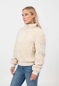 Giacca bomber in pile color crema con colletto alto, zip frontale e polsini a costine. Presenta tasche laterali e una finitura testurizzata.