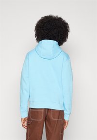 Persona con cabello rizado, vistiendo una sudadera azul clara y pantalones marrones con costuras blancas, de espaldas contra un fondo blanco liso.