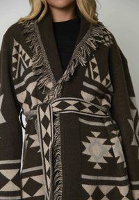 Vrouw in een bruine en beige geometrisch patroon gebreide cardigan met franjes en een bijpassende riem om de taille geknoopt.
